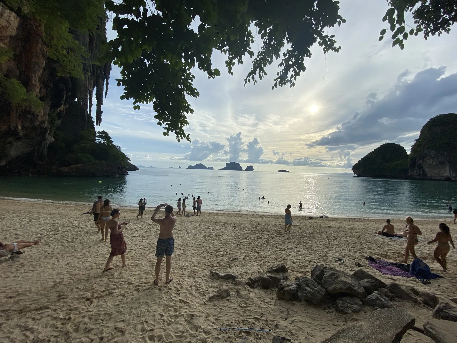 Beachgoers and swimmers beneath cliffs and overhanging branches in Thailand